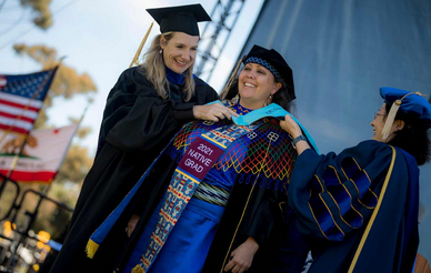 Commencement graphic of two associate deans hooding a Native American doctoral graduate